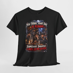 May include: A black t-shirt featuring a graphic print of three witches sitting at a bar with bottles and candles. The text reads: YOU THINK YOUR DAY IS ROUGH... SOMEBODY DROPPED A HOUSE ON OUR SISTER. The scene includes a dark tavern background.