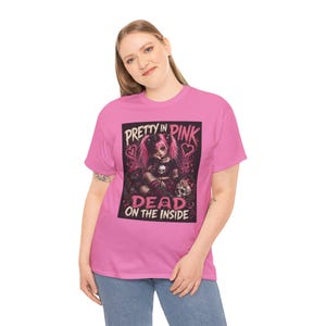 May include: A bright pink t-shirt featuring a graphic print of a character with pink hair, a skull motif, and a teddy bear. The text reads PRETTY IN PINK DEAD ON THE INSIDE in a stylized font, set against a dark rectangular background.
