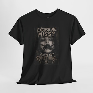 May include: A black short-sleeve t-shirt featuring a graphic print of a person with a large spider over their mouth. The text reads: EXCUSE ME, MISS? YOU'VE GOT SOMETHING... DL XXVIII. The design is in shades of grey, black, and muted brown.