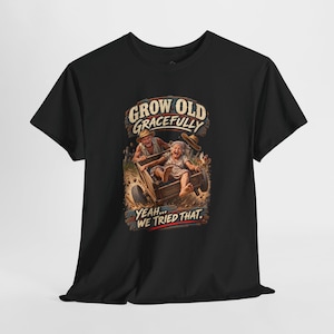 May include: A black t-shirt featuring a graphic of an elderly couple riding in a wooden cart. The text above reads "GROW OLD GRACEFULLY" and the text below reads "YEAH... WE TRIED THAT." The illustration shows the couple laughing with a hat flying.