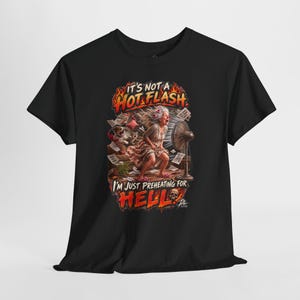 May include: A black short-sleeved t-shirt featuring a graphic of a person standing in front of a fan with the text IT'S NOT A HOT FLASH I'M JUST PREHEATING FOR HELL. The design includes flames, flying papers, a small dog, and a skull illustration.