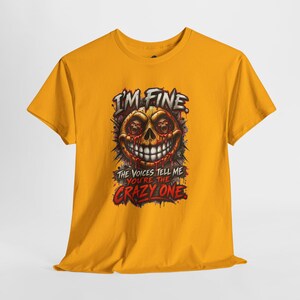 May include: A mustard yellow short-sleeve t-shirt featuring a graphic print of a grinning skull with smaller skulls in its eye sockets. The text reads I'M FINE. THE VOICES TELL ME YOU'RE THE CRAZY ONE. The design includes red and black accents.