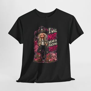 May include: A black t-shirt featuring a graphic of a blonde character in a witch hat, dark jacket, and boots, standing next to a pink dress on a hanger and a pink rabbit toy. The text reads: I WILL NOT WEAR A FUCKING DRESS!!! in bold pink letters.