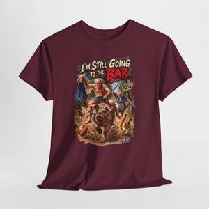 May include: A maroon short-sleeve t-shirt featuring a graphic of an older person riding a pig while holding a beer mug, with another person chasing them with a rolling pin. The text reads "I'M STILL GOING TO THE BAR" above a wooden sign pointing to a bar.