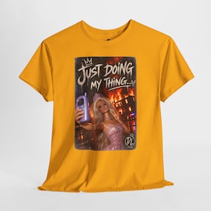 May include: A mustard yellow t-shirt featuring a graphic print of a blonde doll in a pink dress taking a selfie with a purple phone. The background shows a building on fire at night. The text reads JUST DOING MY THING with a crown and a heart.