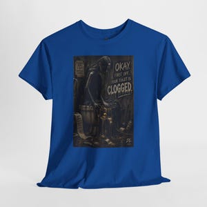 May include: A royal blue short-sleeve t-shirt featuring a dark, detailed graphic print of a long-haired figure sitting on a toilet. The text reads, "OKAY, FIRST OFF... YOUR TOILET IS CLOGGED." The design includes a roll of toilet paper and debris.