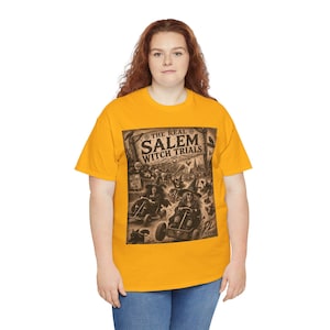 May include: A mustard yellow short-sleeve t-shirt featuring a dark brown graphic print. The design depicts witches in go-karts with the text "THE REAL SALEM WITCH TRIALS 1692" and "GO WITCHES GO!" along with various figures and a small skull icon.