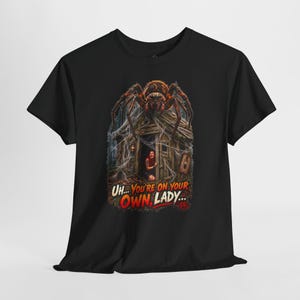 May include: A black t-shirt featuring a graphic print of a large spider perched atop a dilapidated wooden shack covered in cobwebs. A person is visible in the doorway. The text reads, "UH... YOU'RE ON YOUR OWN, LADY..." in orange and white lettering.