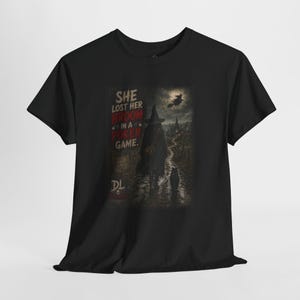 May include: A black short-sleeve t-shirt featuring a graphic print of a witch walking with a cat on a path toward a distant building under a full moon. The text reads: SHE LOST HER BROOM IN A POKER GAME. The initials DL appear in the bottom left corner.