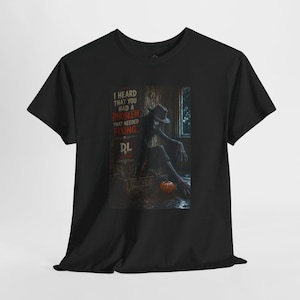 May include: A black short-sleeved t-shirt featuring a graphic print of a dark, shadowy figure in a hat sitting in a room. The text reads: I HEARD THAT YOU HAD A PROBLEM THAT NEEDED FIXING. A small orange pumpkin sits on the floor near the figure.