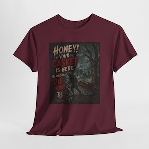 May include: A maroon short-sleeve t-shirt featuring a graphic print of a person carrying a casket in a graveyard. The text reads: HONEY! YOUR CASKET IS HERE! GO AHEAD AND DIE NOW! DL. The design includes dark, moody tones and a gothic atmosphere.