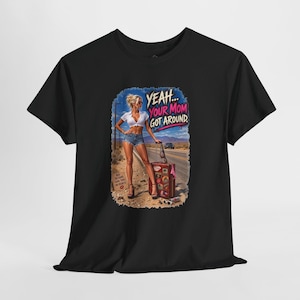 May include: A black short-sleeve t-shirt featuring a graphic print of a person standing on a desert road next to a suitcase. The text reads YEAH... YOUR MOM GOT AROUND. The graphic includes a desert landscape, a road, and a sign with text.