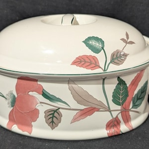 May include: An oval ceramic casserole dish with a matching lid, featuring a cream-coloured base decorated with a botanical pattern of pink flowers and green and brown leaves. The dish includes two side handles and a central lid knob.