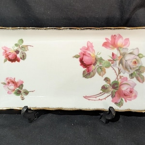 May include: A rectangular white porcelain serving tray with a scalloped edge and a gold-trimmed rim. The surface features a large cluster of pink and white roses on the right side and two smaller individual pink rose blossoms on the left side.