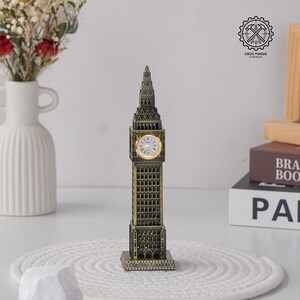 Puede incluir: Un modelo de metal color bronce de la torre del reloj Big Ben con detalles arquitectónicos intrincados, una aguja puntiaguda y una esfera de reloj circular con manecillas y números dorados sobre una base rectangular.