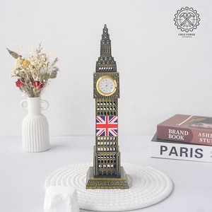 May include: A bronze-coloured metal model of the Big Ben clock tower featuring a circular analogue clock face with black hands and numerals, a Union Jack flag graphic on the front, and the text Big Ben London embossed on the base of the structure.