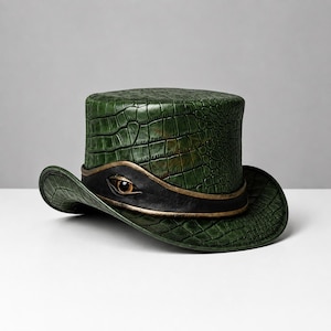 May include: A green top hat featuring a textured crocodile skin pattern. The hat is accented with a black leather band that wraps around the base, featuring a single realistic eye detail centred at the front with metallic gold trim edges.