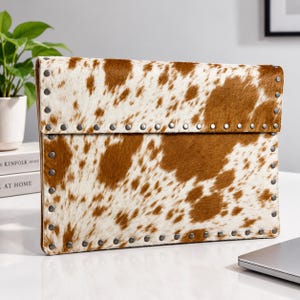 May include: A rectangular clutch bag featuring a brown and white cowhide pattern. The exterior is accented with a border of small, metallic silver-toned studs along the edges and the flap closure, providing a structured and detailed appearance.