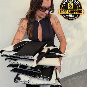 Puede incluir: Una pila de bolsos de mano rectangulares hechos de piel de vaca en blanco y negro. Cada bolso presenta un patrón de manchas de vaca distinto y está decorado con una fila de tachuelas de metal plateado en los bordes. Diseño plano y estructurado.