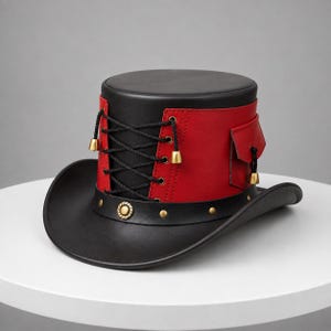 May include: A black and red top hat featuring a black lace-up corset detail on the side, a small side pocket with a flap, and a black hatband adorned with gold-toned metal studs and a central circular emblem with a rope-like texture design.