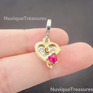 May include: A two-tone metal charm pendant featuring a silver-coloured heart base with the engraved text YOU ARE THE ONE and a gold-coloured heart overlay accented with a small, bright pink heart-shaped stone at the bottom centre.