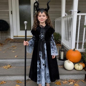 May include: A black velvet dress with long flared sleeves and a grey patterned skirt, featuring a lace-up bodice. The outfit includes a black headband with two curved horns and a tall black staff topped with a silver-colored skull ornament.