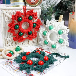 May include: Three macrame snowflake ornaments in red, white, and dark green, each featuring six round wooden beads. The red and white ornaments are hung on a macrame container, while the green one rests on a printed tutorial instruction sheet.