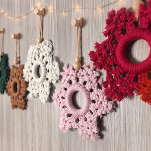 May include: A series of macrame snowflake ornaments in dark green, brown, cream, light pink, and red, each featuring a central ring, knotted cord details, and a wooden bead, suspended from individual light-colored wooden wall hooks.