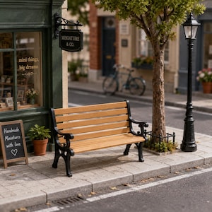 Puede incluir: Un banco de parque en miniatura con listones de madera de color marrón claro y reposabrazos y patas de metal negro con volutas. El banco está situado en una acera pavimentada junto a un escaparate verde con un letrero Miniature & Co. y una farola negra.