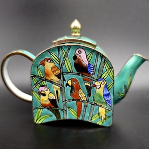 May include: A small, turquoise-coloured teapot featuring a decorative enamel design of five colourful parrots perched on bamboo branches. The teapot has a gold-toned metal handle, spout, and a small, pointed lid finial, with a flat, arched base.