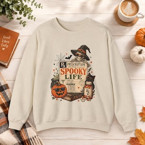 May include: A beige crewneck sweatshirt featuring a graphic print of a skeleton wearing a witch hat and sunglasses, a jack-o'-lantern, a black cat, and a prescription bottle. The text reads Prescription for a Spooky Life with checkboxes.