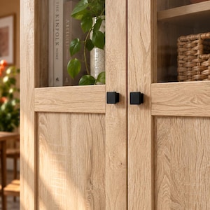 May include: A light oak wood cabinet featuring two glass-panelled doors with small, square, matte black metal knobs. The cabinet interior displays a book titled The Kinfolk Home, a green leafy plant in a white vase, and a woven basket on a shelf.