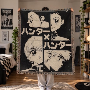 May include: A black and cream woven throw blanket featuring a grid layout of four anime character portraits and the Japanese text "ハンター×ハンター" in the center. The blanket has a fringed edge and a textured, tapestry-style construction.