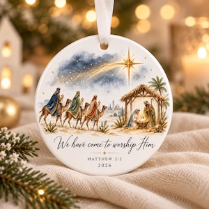 May include: A round white ceramic Christmas ornament featuring a watercolour illustration of the Nativity scene with three wise men on camels, a stable, and a bright star. The text reads We have come to worship Him, MATTHEW 2:2, and 2026.