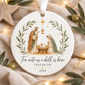 Peut inclure: Une décoration ronde en céramique blanche illustrant une scène de nativité avec des personnages en robes marron et vertes, une crèche et une lanterne suspendue. Le texte indique "For unto us a child is born", "ISAIAH 9:6" et "2026".