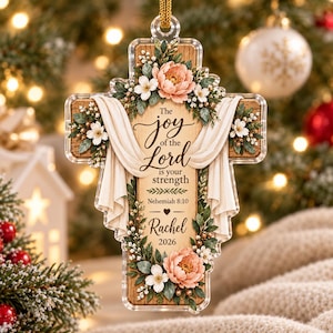 May include: A cross-shaped acrylic ornament featuring a wood-grain background, decorated with peach-coloured peonies and white flowers. The text reads: The joy of the Lord is your strength, Nehemiah 8:10, Rachel, 2026. Draped white fabric accents the design.