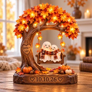 May include: A decorative tabletop light featuring a brown tree with orange and red autumn leaves, illuminated by small lights. Two white ghost figures wearing a plaid scarf sit on a swing. The tree trunk has a heart with the text Emma + Levi, and the base displays the date 10.10.26 with two hearts. Small pumpkins and acorns are at the base.