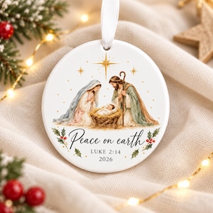 Peut inclure: Une décoration de Noël ronde en céramique blanche illustrant une scène de nativité avec Marie, Joseph et l'enfant Jésus dans une crèche. Une étoile dorée brille au-dessus. Le texte Peace on earth, LUKE 2:14 et 2026 est inscrit en dessous.