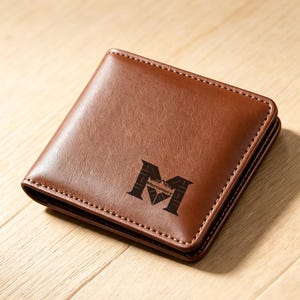 May include: A brown bi-fold wallet with a smooth, textured surface and visible stitching along the edges. The front features a dark, engraved logo consisting of a large letter M with the word Mercenary printed inside the center of the letter.