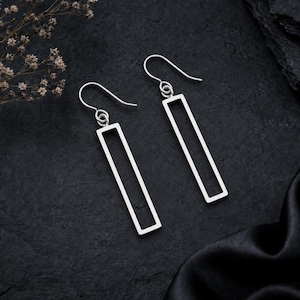 May include: A pair of silver-toned earrings featuring an open rectangular frame design. Each earring hangs from a curved hook wire, showcasing a clean, geometric silhouette with a smooth metallic finish that reflects light against the dark background.