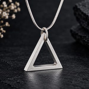 Op de afbeelding: Een zilverkleurige ketting met een holle, driehoekige hanger aan een fijne slangenketting. De hanger heeft een geborstelde matte afwerking en is met een kleine ronde ring aan de ketting bevestigd, wat zorgt voor een geometrisch ontwerp.
