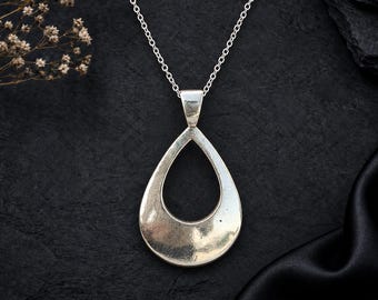 Puede incluir: Un collar de tono plateado con un gran colgante en forma de lágrima abierta suspendido de una cadena fina. El colgante tiene una superficie lisa y reflectante con un acabado ligeramente desgastado, creando un diseño sencillo.