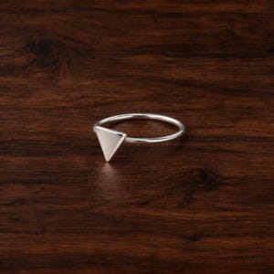 May include: A thin, polished silver-tone ring featuring a small, flat, equilateral triangle charm attached to the top of the circular band. The metal surface is smooth and reflective, catching light to highlight the geometric shape of the ring.