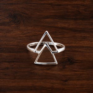 Peut inclure: Une bague en argent poli présentant un motif géométrique composé de deux triangles ouverts superposés. L'anneau est lisse et circulaire, soutenant la structure triangulaire qui s'étend vers le haut depuis le sommet de la bande principale.