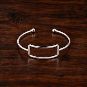 Può includere: Un bracciale aperto color argento caratterizzato da un design a cornice rettangolare centrale. Il bracciale presenta una finitura liscia e lucida e termina su entrambe le estremità con piccoli accenti sferici, creando un'estetica geometrica pulita.