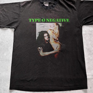 以下が含まれることがあります： 鮮やかな緑色の文字でTYPE O NEGATIVEと書かれた黒の半袖Tシャツ。長い黒髪の人物が長い金髪の人物を抱きしめているグラフィックプリントが施されている。Tシャツはクルーネックで、襟元にタグが付いている。