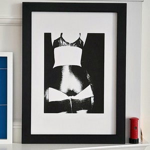 Puede incluir: Fotografía en blanco y negro del torso de una persona, que lleva un sujetador negro y ropa interior blanca. La foto está enmarcada en un marco negro.