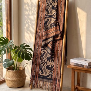 Puede incluir: Una manta textil rectangular con una base negra oscura y complejos patrones geométricos y motivos de aves estilizadas en tonos beige y naranja. La tela presenta bordes con rayas verticales y un borde con flecos en la parte inferior, sobre una escalera.
