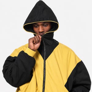 Può includere: Una giacca con cappuccio nera e giallo senape dotata di cerniera frontale a tutta lunghezza, collo alto e polsini a coste. Il capo presenta un design a blocchi di colore con maniche nere e corpo giallo senape, accentuato da profili gialli lungo il cappuccio.