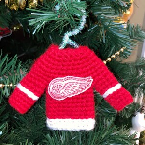May include: A red crocheted sweater ornament with a white wing logo, hanging on a Christmas tree.
